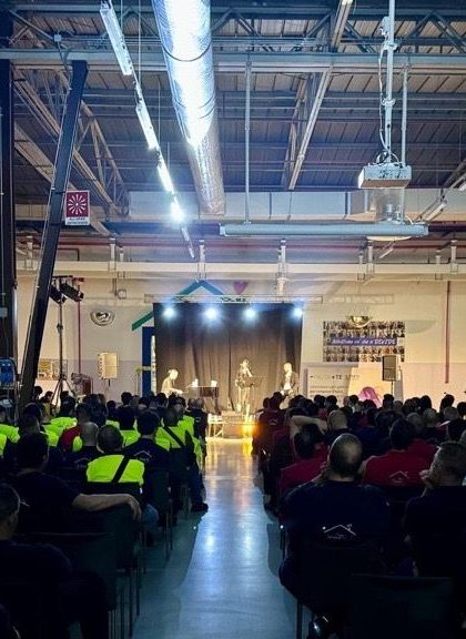 Un grande gruppo di persone sedute in un capannone assiste a una presentazione su un palco illuminato.