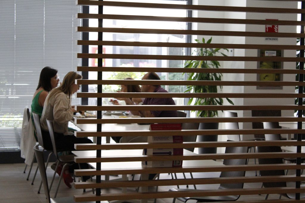 Quattro persone sedute a un tavolo di una mensa mentre discutono, viste attraverso una parete con listelli di legno orizzontali.