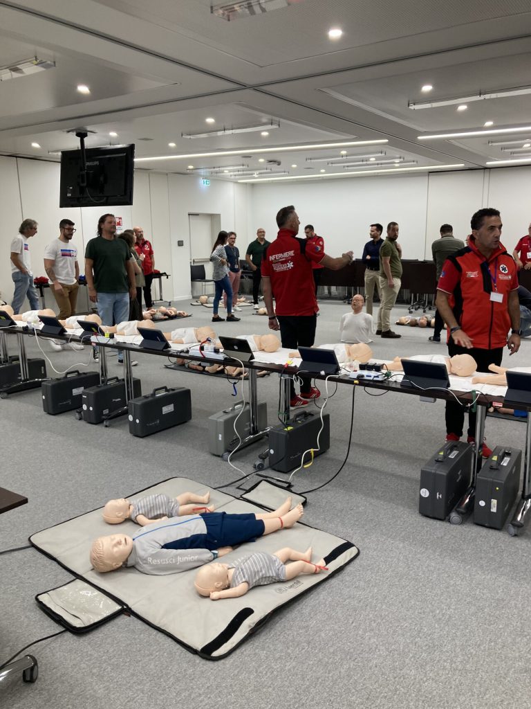 In una sala corsi, diversi manichini per esercitazioni di primo soccorso sono disposti a terra e sulle panche, mentre istruttori e partecipanti si preparano all’attività.