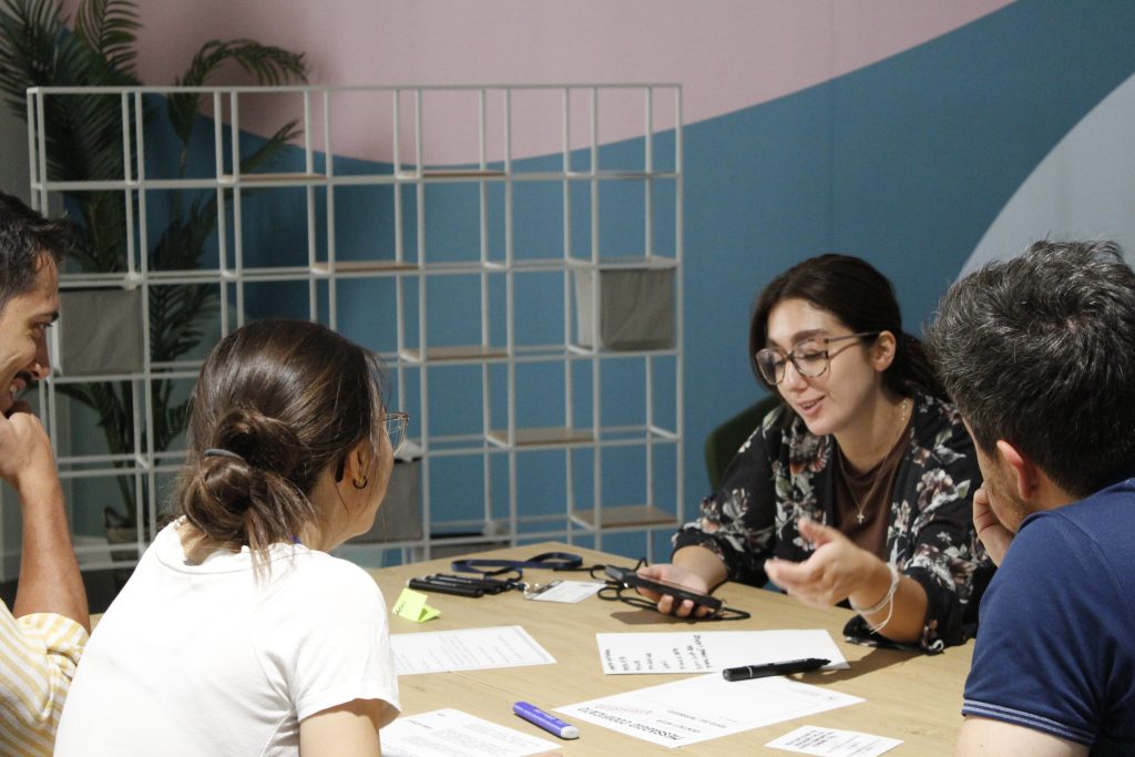 A woman smiles while talking with a colleague sitting across from her, as they discuss some papers and notes on a table.