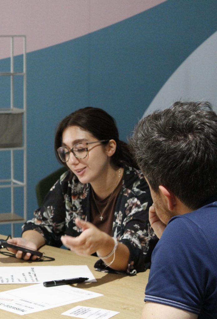 Ragazza parla sorridendo con un collega seduto di fronte, mentre discutono su alcuni fogli e appunti su un tavolo.