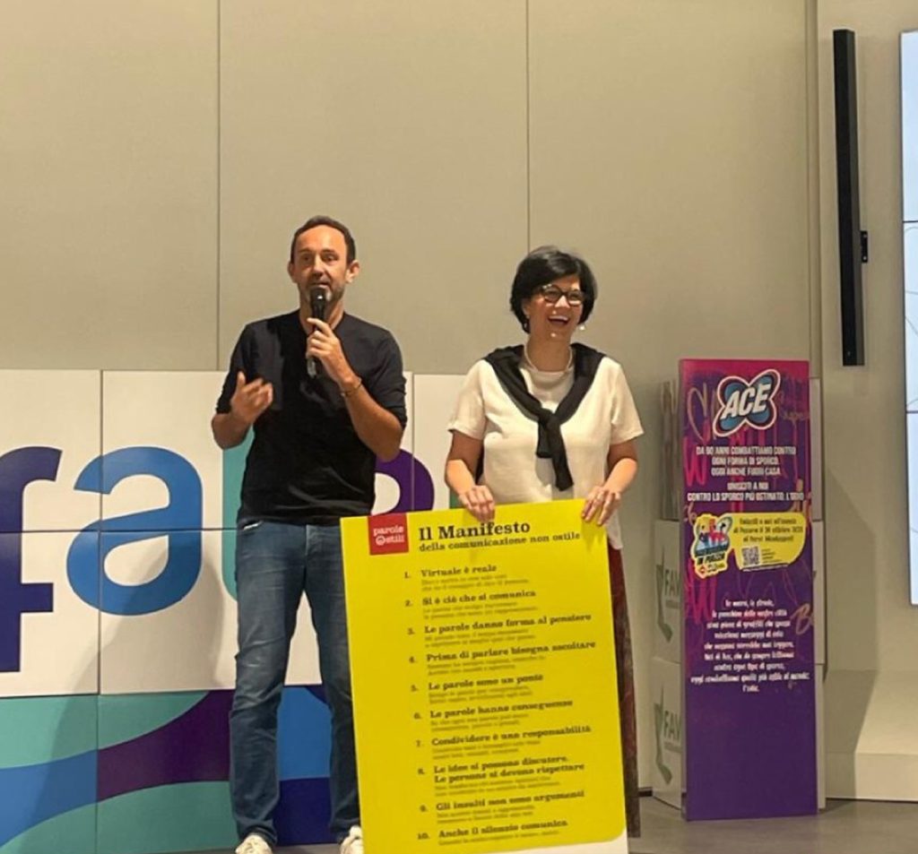 Un uomo con microfono e una donna accanto a un grande cartellone giallo del Manifesto ACE parlano a un evento.