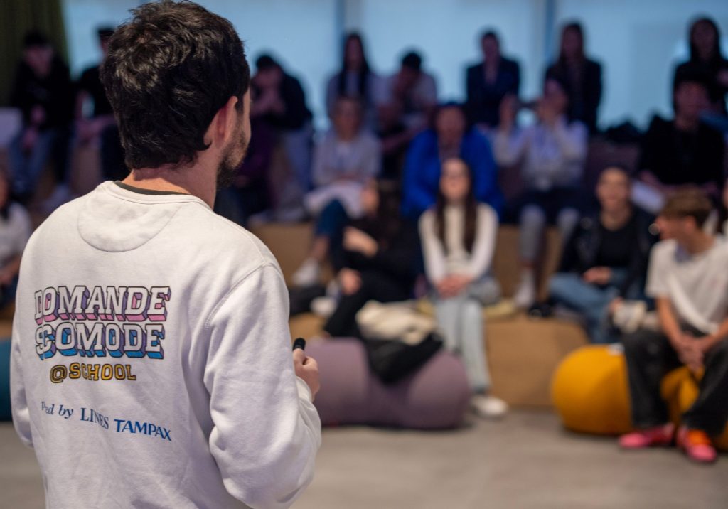 Un ragazzo di spalle con una felpa con la scritta Domande Scomode @School parla davanti a un gruppo di studenti seduti.