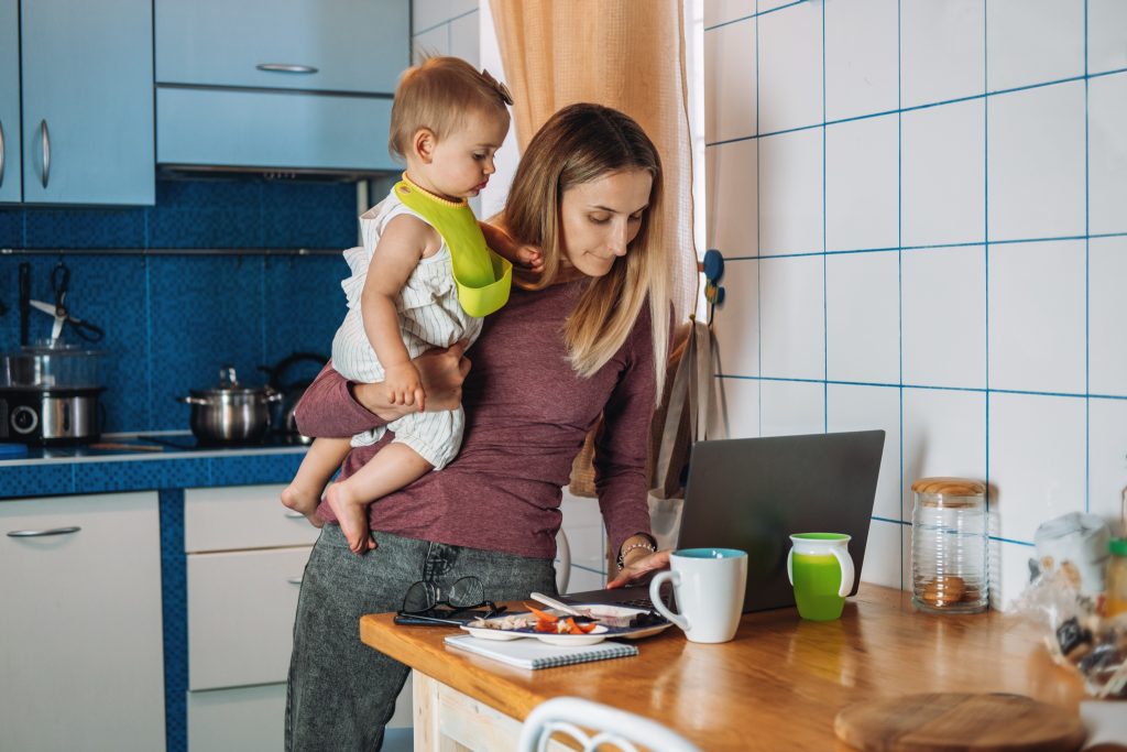 Mamma con in braccio una bambina lavora al computer in cucina con tazza e quaderno sul tavolo.