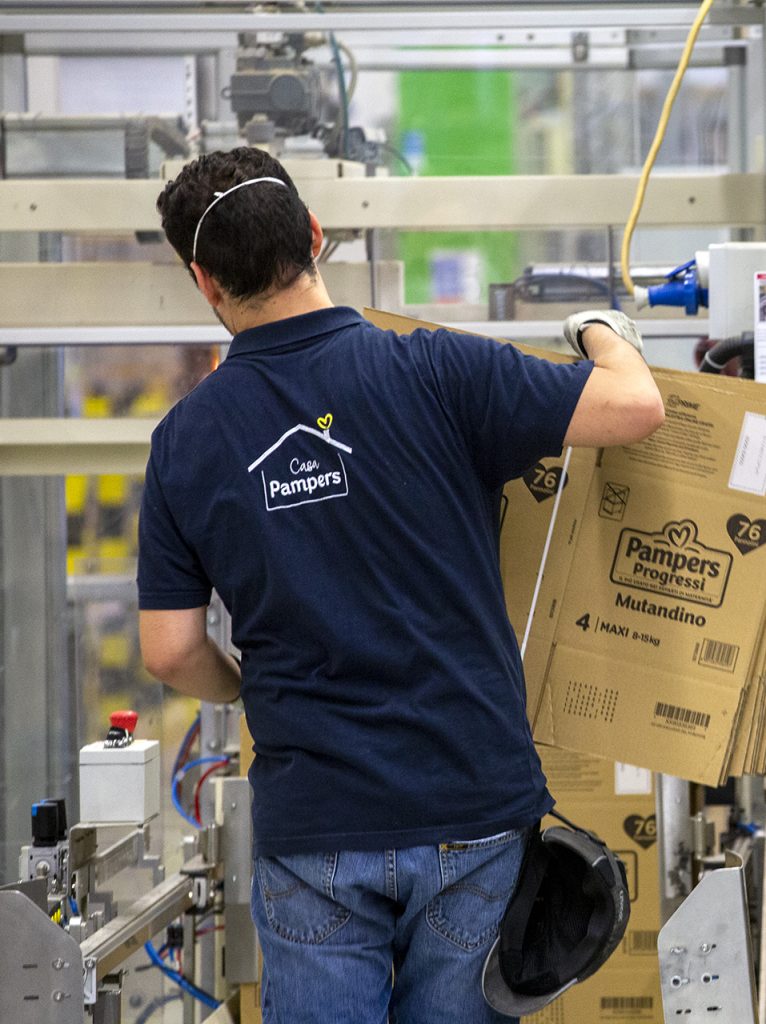Operaio con maglietta aziendale lavora accanto a un macchinario industriale, spostando scatole di cartone.