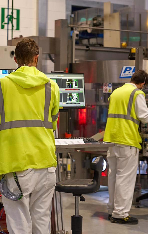 Due operatori in giacca gialla ad alta visibilità e pantaloni bianchi lavorano in un impianto industriale, uno davanti a un computer e l’altro su un macchinario di produzione.