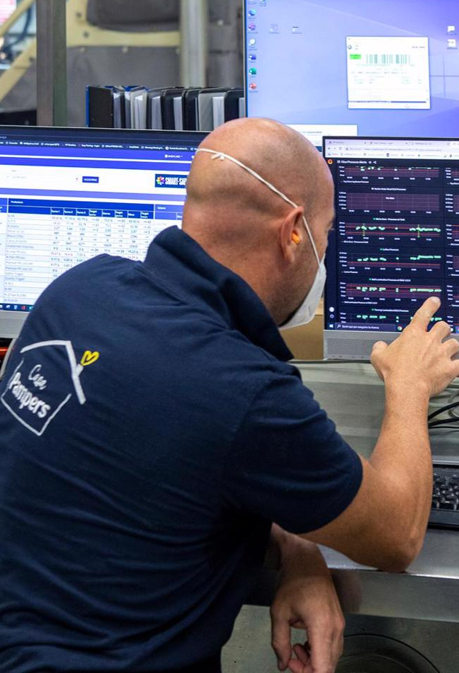 Operaio con maglietta aziendale controlla i dati su più monitor in fabbrica.