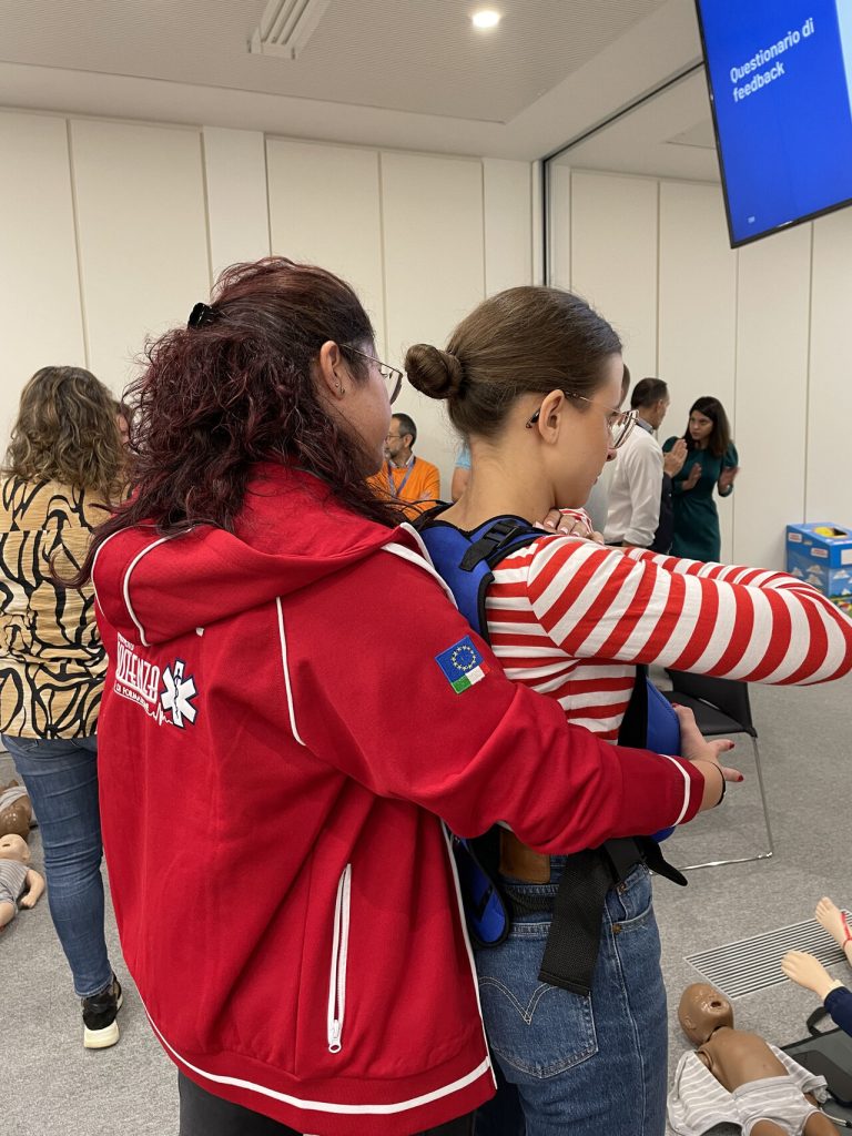 First aid instructor wearing a red jacket guides a young participant during a practical training exercise.