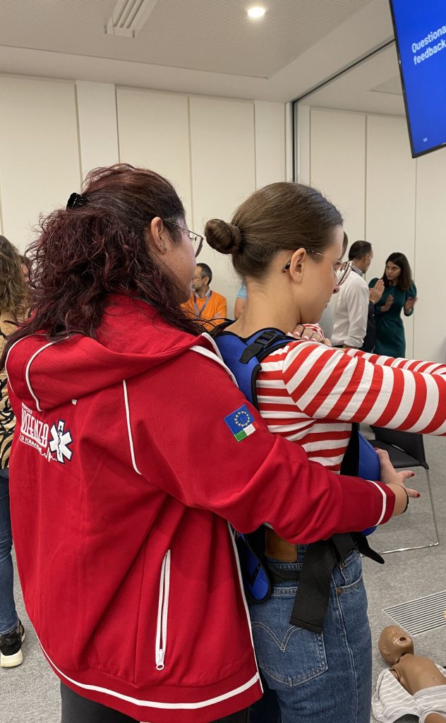 Istruttrice di primo soccorso in giacca rossa guida una giovane partecipante durante un’esercitazione pratica.