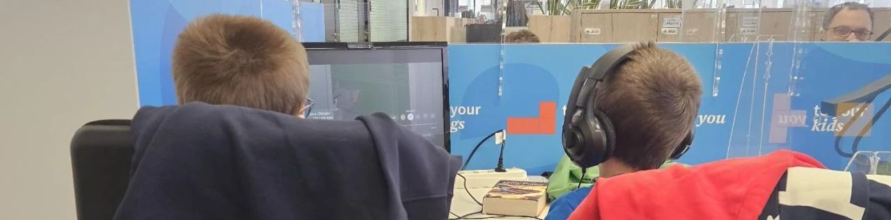 Due bambini di spalle davanti a uno schermo, uno indossa le cuffie mentre giocano al computer.